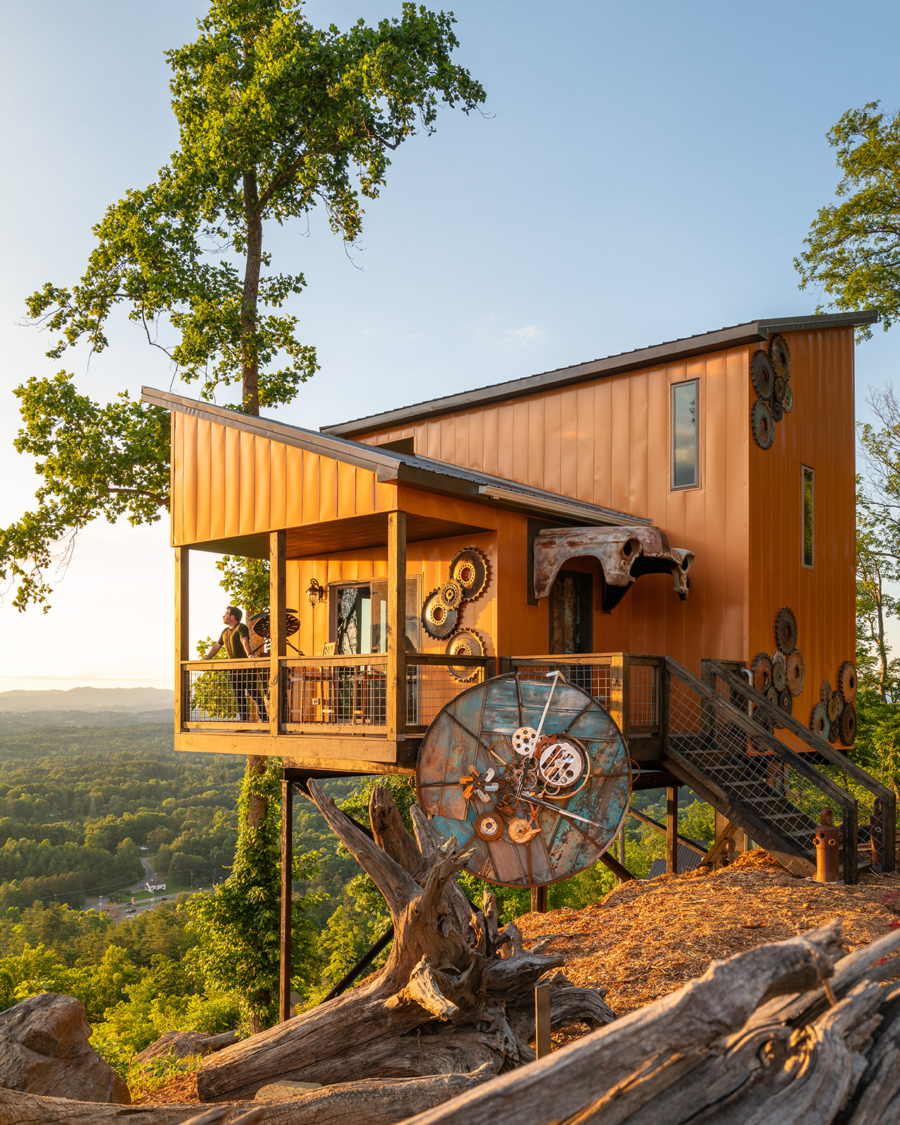 Copper-colored Max dwelling with decorative gears and a large metal wheel, overlooking wooded mountains.